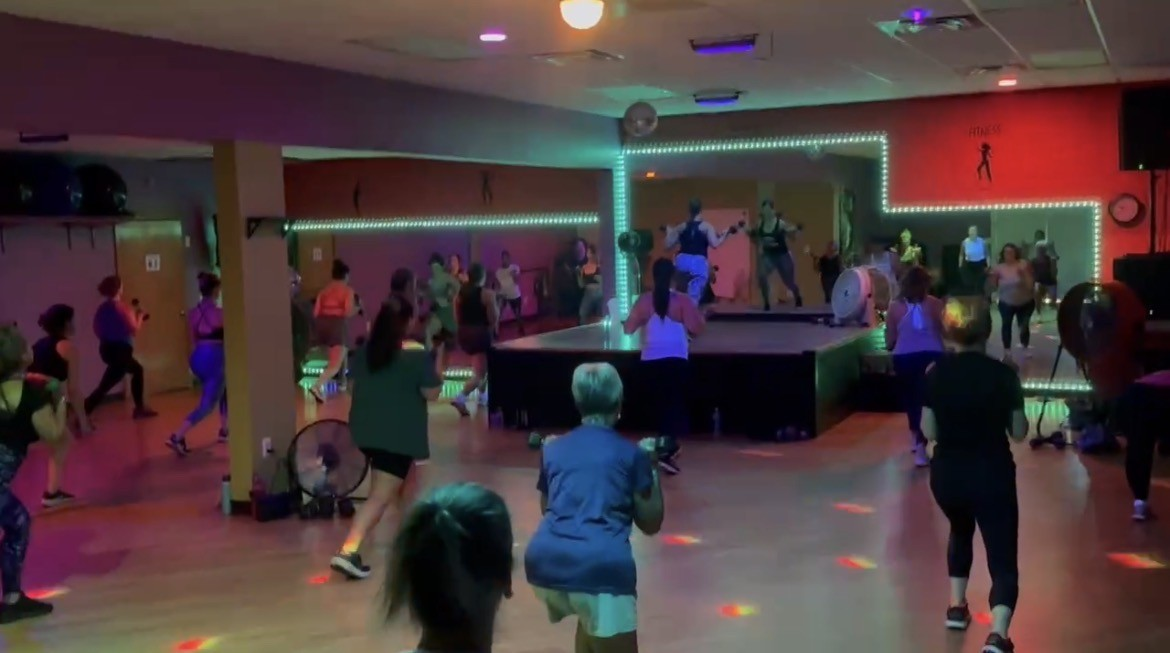 Group fitness class training on the studio floor with LED-lit stage and colorful lighting